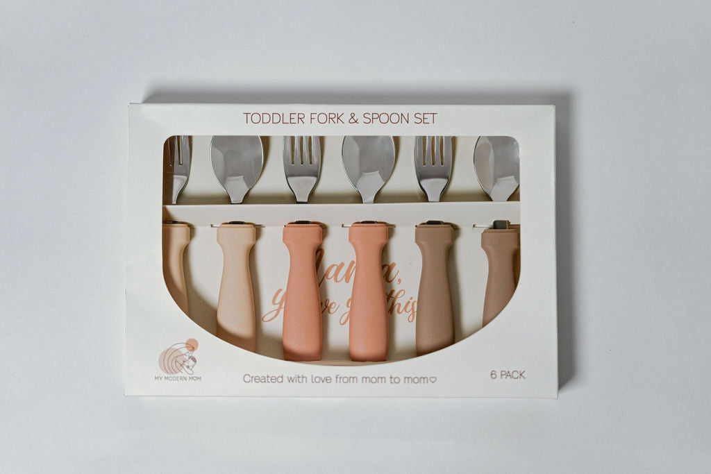 A packaged toddler fork and spoon set with multiple utensils visible through the clear plastic window, primarily in beige and white colors.

