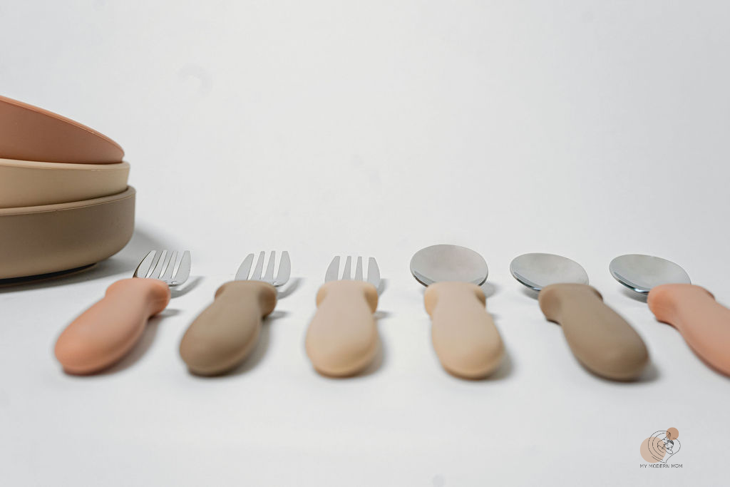 A series of utensils with varying shades of skin tone-colored handles, displayed in a line with spoons on one side and forks on the other, against a white background with a bowl at the end.

