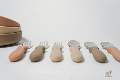 A series of utensils with varying shades of skin tone-colored handles, displayed in a line with spoons on one side and forks on the other, against a white background with a bowl at the end.


